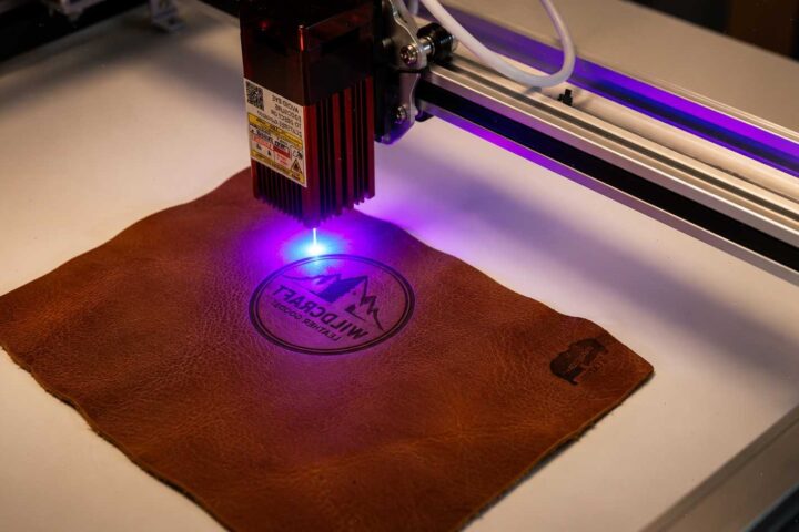 A laser engraver using a purple beam to etch a circular mountain logo onto a piece of brown leather