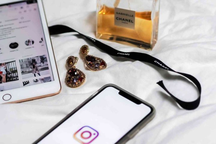 Flat lay of an iPad and smartphone showing Instagram, Chanel perfume, and jewelled earrings on white bedding