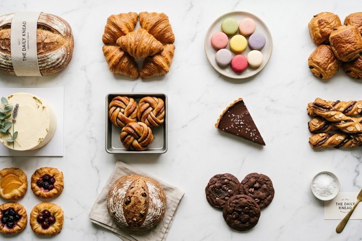 Selection of The Daily Knead bakery products including bread, croissants, macarons, pastries, and cakes on marble